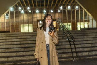 Young woman in a trench coat smiling and looking at her smartphone while standing on modern urban