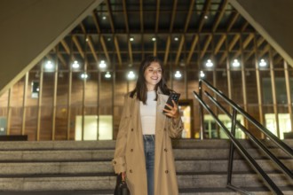 Young woman smiling while checking her smartphone, walking down urban stairs at night, wearing a
