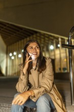 Young woman sitting outdoors at night, smiling brightly while having a phone conversation,