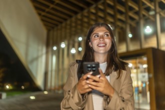 Young woman smiling while holding a smartphone and looking up, exploring a modern city street at