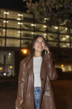 Young woman in a brown leather jacket smiling, talking on a mobile phone in an urban street at