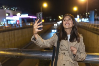 Young woman smiling and holding a smartphone, capturing a selfie against the bright, blurred lights