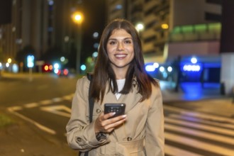 Woman smiling brightly and holding a smartphone, standing on a bustling city street at night with