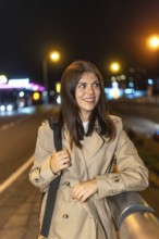Woman smiling on a nighttime city street, wearing a trench coat, looking away as colorful urban