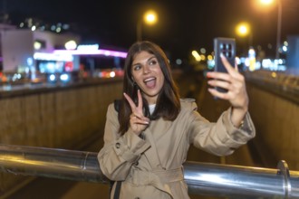 Young woman wearing a trench coat standing on a bridge, holding a smartphone and taking a self
