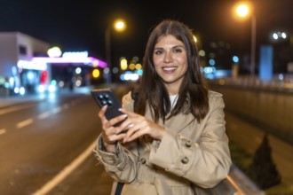 Young woman in a trench coat standing on a city street at night, smiling at her smartphone as she