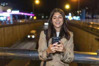 Young woman standing on a bridge at night, smiling as she uses her smartphone with city lights and