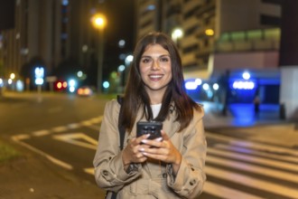 Young woman smiling and interacting with her smartphone while standing on a city street at night,
