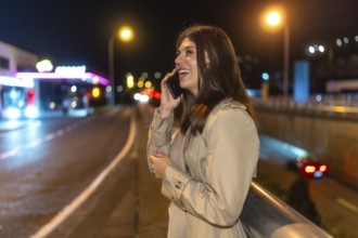 Happy young woman smiling and talking on a mobile phone while standing on an urban street at night,