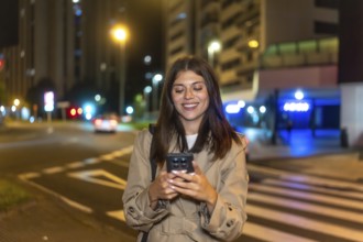 Young woman smiling with eyes closed, enjoying her smartphone on a city street at night, relaxed