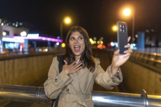 Young woman in trench coat surprised and joyful taking a nighttime selfie on a bustling city