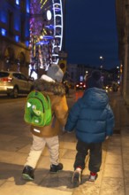 Two children holding hands and walking away from the camera on a sidewalk near a brightly lit