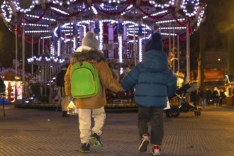 Two young brothers, holding hands, walk away from a brightly lit carousel at a christmas market in
