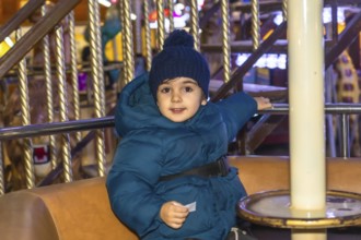 Young child wearing a blue winter coat and hat enjoying a carousel ride at a christmas market in
