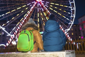 Two children with winter coats and backpacks admiring a brightly lit ferris wheel at a christmas