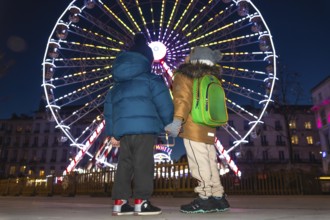 Two children holding hands are admiring a brightly lit ferris wheel at a christmas market in