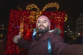 Tourist taking a selfie and making thumbs up sign with christmas lights and decorations in bayonne,