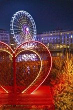 Illuminated heart shaped bench with small christmas trees and ferris wheel in the background,