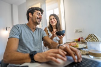Young couple enjoying breakfast in a bright kitchen, working from home on a laptop while sharing