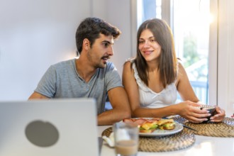 Young couple talking and having breakfast in their kitchen, with a laptop on the table and natural