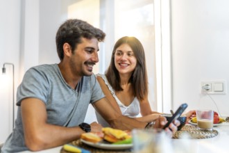 Young couple smiling and enjoying breakfast together in their cozy kitchen, using a smartphone to