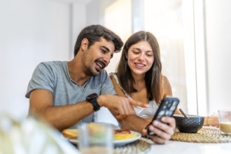 Young couple enjoying breakfast at home, smiling and pointing at a smartphone while browsing social