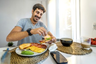 Young man smiling while preparing a delicious sandwich for breakfast at home, enjoying a healthy