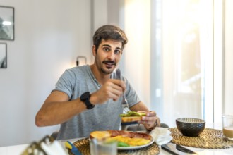 Happy man enjoying avocado toast alongside ham and eggs while relaxing in his modern apartment,