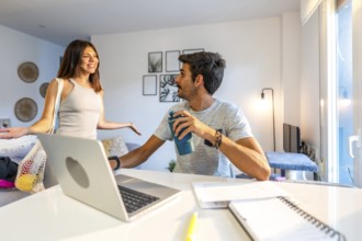 Young woman arriving home with groceries in reusable bag, greeting her boyfriend working remotely