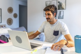Young male freelancer working remotely at home, using a laptop and reviewing notes while sitting