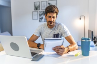 Young man managing his business remotely, reviewing notes and working on his laptop in his bright