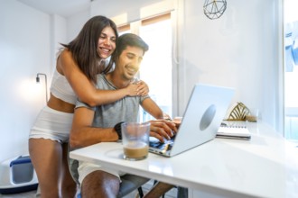 Young smiling couple using laptop at home, woman hugging man working on computer, modern bright