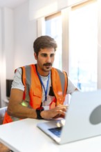 Focused engineer wearing high visibility vest working on laptop in bright modern office, managing