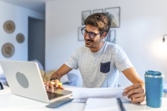 Young man working comfortably at home, engaging with a laptop and reviewing documents, smiling