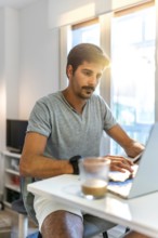 Young man concentrating on his laptop, working remotely from his bright home office while enjoying