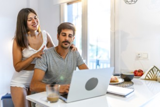 Young woman massaging her husband's shoulders while he's working from home on his laptop in the