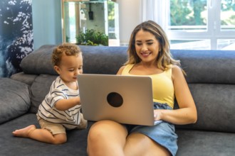 Young mother balancing work and family life, working on a laptop while cuddling her baby son on the