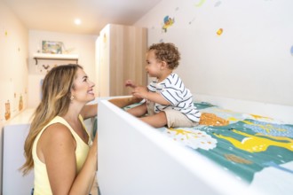Smiling mother looking at her cute baby sitting on a bunk bed in a decorated bedroom, happy family