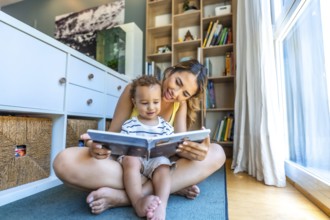 Happy mother spending quality time reading a book to her baby son, sitting on the floor in the