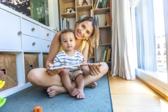Happy mother and her baby son reading a book together, sitting on the floor in the living room at