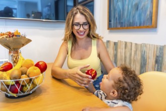 Loving mother giving a fresh peach to her baby son while sitting at the kitchen table, promoting
