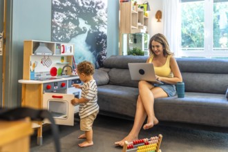 Woman working remotely on laptop while her toddler plays with a toy kitchen, demonstrating work
