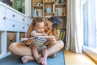 Mom kissing her little son while reading a book together, sitting on the floor in the living room