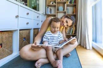 Young mother sitting on the floor reading a book with her little son, enjoying quality time