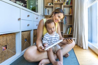 Happy mother and baby son reading a book together, sitting on the floor in the living room at home