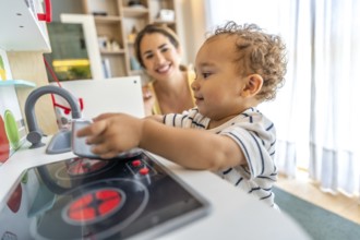 Happy toddler playing with toy kitchen while his mother watches him, promoting early childhood