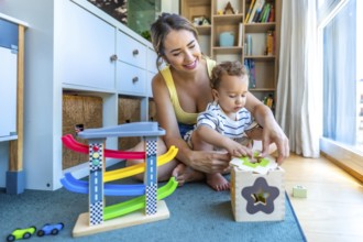 Happy mother helping her baby son playing with a shape sorter toy, sitting on a play mat in a