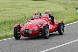 An open red sports car with two drivers drives through a green landscape, classic car, car race,