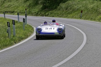 A silver racing car with two people drives swiftly along a winding road, classic car, car race,
