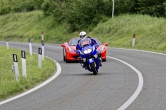 Blue motorbike takes a bend in front of a red Ferrari convertible on a country road, classic car,